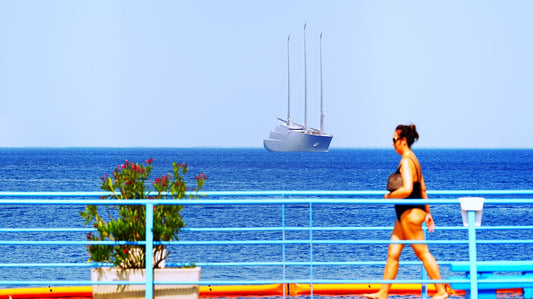 Woman walks along a railing with a large sailboat offshore.