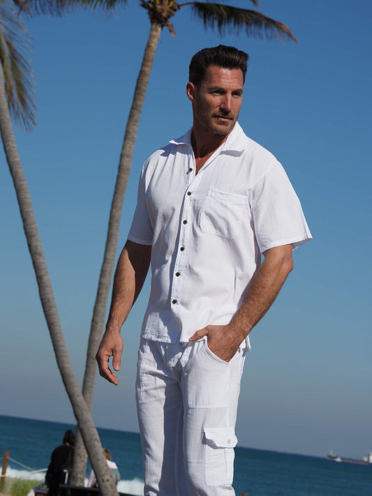 Man in a white outfit standing on a beach with palm trees and blue sky.