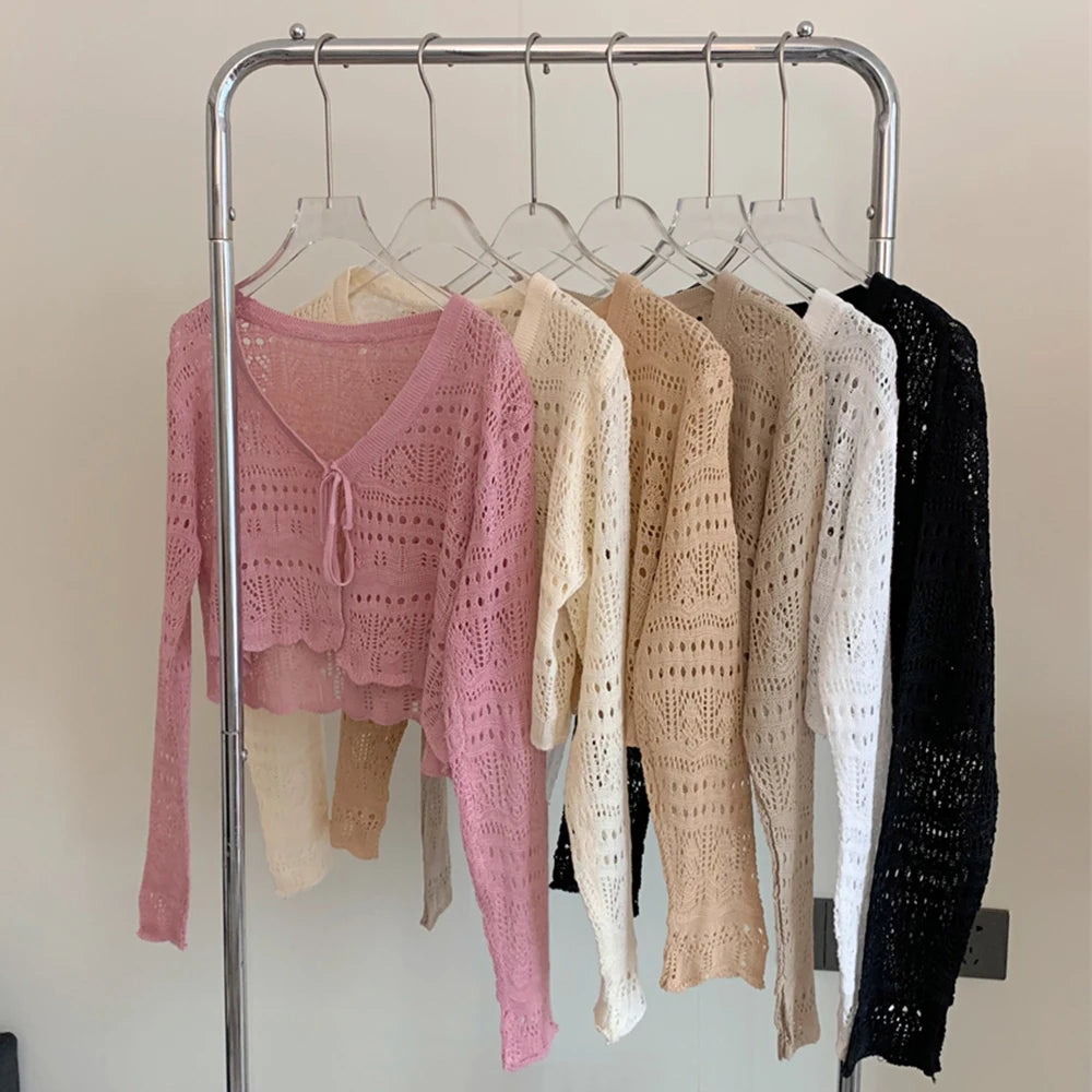Six open-knit long-sleeve tops in different colors hanging on a metal clothing rack against a plain light wall. The front top is pink, with cream, beige, light beige, white, and black tops behind it.