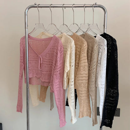 Six open-knit long-sleeve tops in different colors hanging on a metal clothing rack against a plain light wall. The front top is pink, with cream, beige, light beige, white, and black tops behind it.