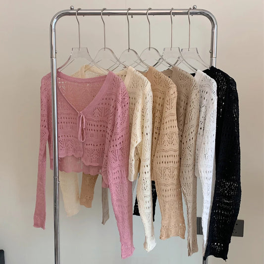 Six open-knit long-sleeve tops in different colors hanging on a metal clothing rack against a plain light wall. The front top is pink, with cream, beige, light beige, white, and black tops behind it.
