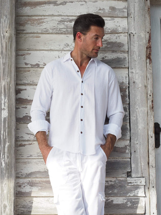 Man wearing a white shirt and pants standing against a rustic wooden wall.